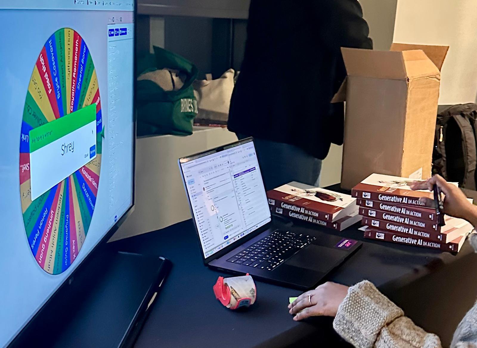 A raffle wheel of entrant names spinning on a large screen at the Anaconda booth, next to a laptop and stacks of Generative AI in Action books